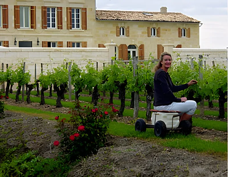 Une femme travaillant dans le vignoble à l'aide d'un petit chariot utilitaire tout en taillant les vignes, avec le bâtiment du château visible en arrière-plan.