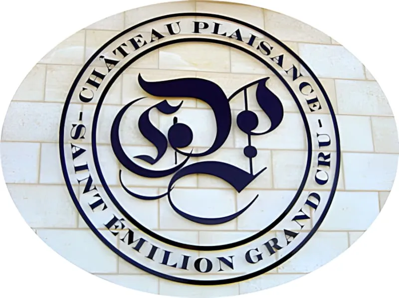 Un logo métallique circulaire monté sur un mur de pierre blanche avec les initiales ornées « CP » en écriture gothique et le texte « Château Plaisance » et « Saint-Émilion Grand Cru » autour de la bordure.