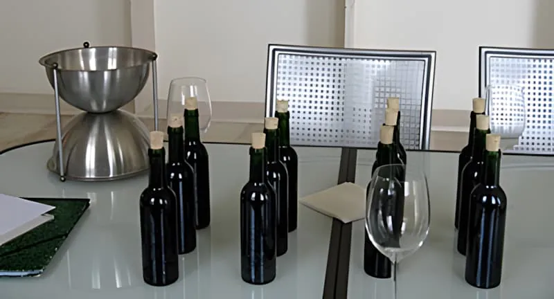 Une installation de dégustation de vin formelle avec plusieurs bouteilles de vin sombres, un crachoir en argent, et des verres à vin disposés sur une table.
