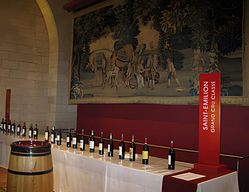 Une longue table bordée de nombreuses bouteilles de vin rouge, installée pour un événement de dégustation professionnel dans une salle avec une grande tapisserie.
