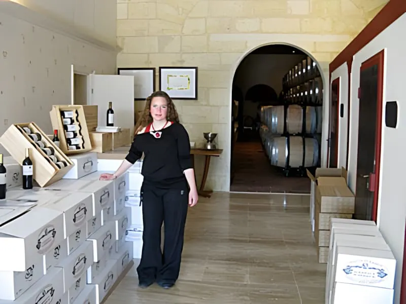 Une jeune femme en vêtements sombres debout dans une salle de dégustation avec des caisses de vin et une vue sur le chai à barriques derrière elle.