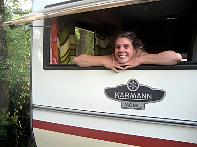 Une jeune femme souriante aux cheveux blonds se penchant par ce qui semble être une fenêtre de maison mobile ou caravane.
