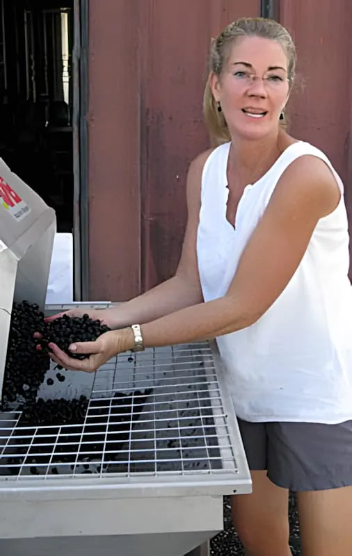 Une femme blonde en débardeur blanc souriant tout en triant des raisins foncés sur une table de tri devant un bâtiment.