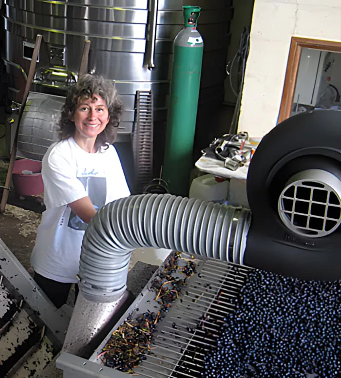 Une femme aux cheveux bouclés souriant tout en travaillant à une station de tri de raisins entourée d'équipements de vinification dans une cave.