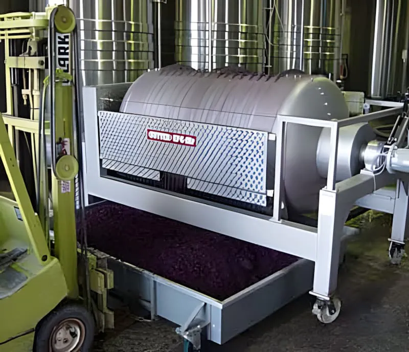 Une grande machine de pressoir cylindrique dans une cave, avec du vin sombre ou du marc de raisin visible, utilisée pour séparer les peaux et pépins du vin.