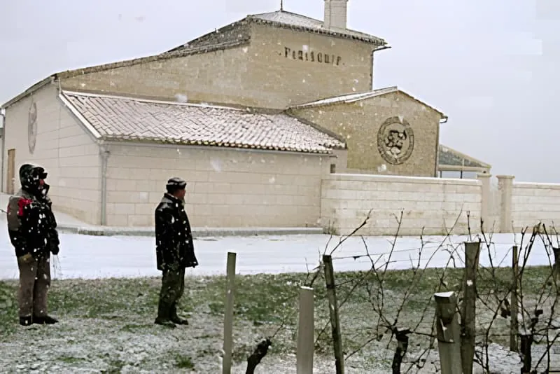 Deux ouvriers en vêtements d'hiver sombres debout près d'un bâtiment de cave en pierre pendant de fortes chutes de neige, avec des rangs de vignoble nus visibles au premier plan.