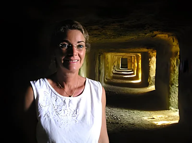 Une femme en blanc debout dans un tunnel en calcaire sculpté avec des murs de couleur ocre chaude, avec des chambres supplémentaires visibles s'étendant au loin.