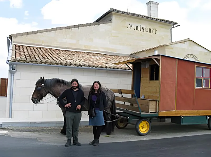 Deux personnes debout près d'un cheval et d'un wagon de voyage coloré devant un bâtiment blanc avec 'Plaisance' écrit dessus.
