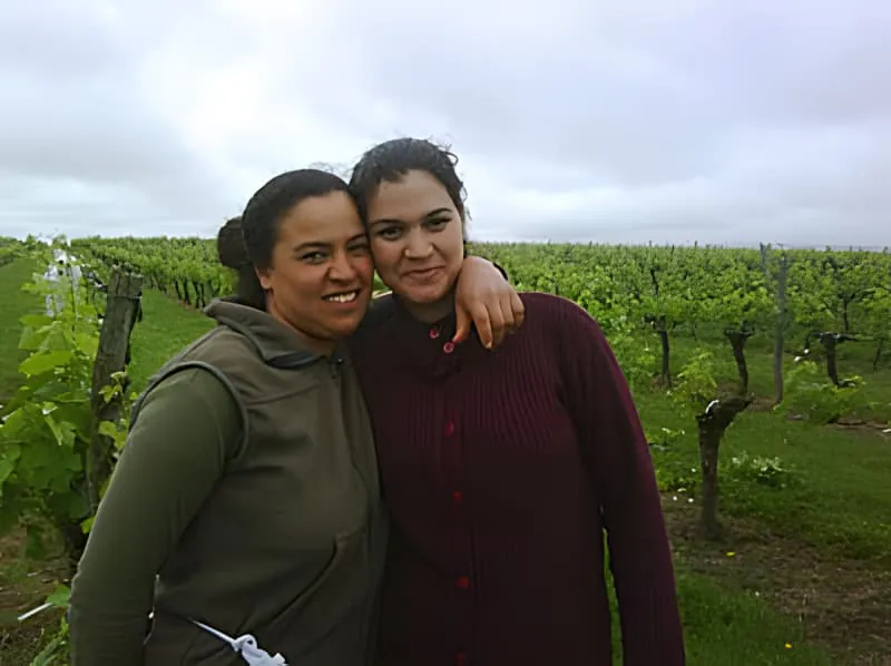 Deux femmes souriantes debout l'une près de l'autre dans un vignoble, l'une portant une veste de couleur claire et l'autre des vêtements plus sombres.