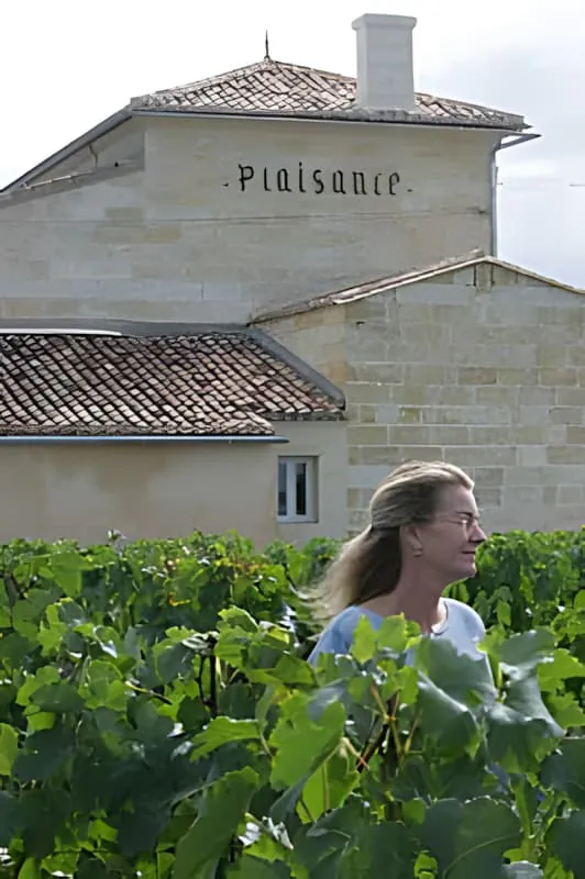Une femme aux cheveux blonds travaillant parmi des vignes vertes luxuriantes avec le bâtiment en pierre blanche du Château Plaisance visible en arrière-plan.